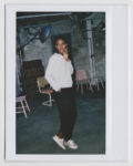 Une personne se tient debout dans une pose détendue, regardant sur le côté, une main touchant son visage et l'autre dans sa poche. Elle porte un haut blanc à manches longues, un pantalon foncé et des baskets blanches, dégageant une ambiance mode parisienne dans une pièce de style industriel avec des chaises et des équipements dispersés.