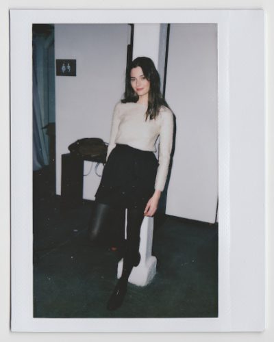Une mannequin aux cheveux longs est appuyée contre un pilier blanc, magnifiquement capturée par le photographe Thomas Duclert. Elle porte un pull clair, une jupe foncée et des collants foncés. Dans l'arrière-plan faiblement éclairé de cette image de style Polaroïd prise à Paris, on peut voir un panneau indiquant les toilettes et quelques meubles.