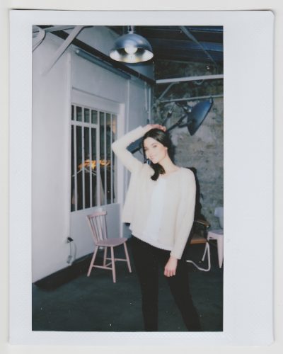 Une personne se tient debout dans une pièce, posant avec une main derrière la tête. Elle porte un haut blanc et un pantalon noir. Il y a une chaise rose, une table avec des objets dessus et un luminaire suspendu en arrière-plan. L'image a l'apparence d'une photo instantanée du photographe Thomas Duclert.