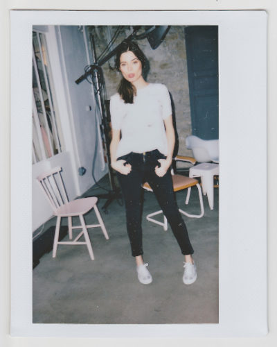Une mannequin se tient à l'intérieur dans une pose décontractée, habillée de façon décontractée d'un t-shirt blanc, d'un jean noir et de baskets blanches. Ses mains sont dans ses poches. L'arrière-plan comprend des chaises, une porte et une fenêtre. La configuration de l'éclairage rehausse la scène avec un air de mode parisienne. L'image a une bordure de type Polaroïd.