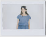 Une mannequin aux cheveux longs et bruns porte un bandeau bleu et une élégante robe en jean bleu. Elle sourit et se tient debout sur un fond blanc uni, reflétant la mode parisienne. L'image a une bordure de style Polaroïd qui lui donne une ambiance chic et sans effort rappelant le travail de Thomas Duclert.