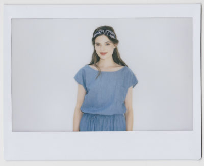 Une mannequin aux cheveux longs et bruns porte un bandeau bleu et une élégante robe en jean bleu. Elle sourit et se tient debout sur un fond blanc uni, reflétant la mode parisienne. L'image a une bordure de style Polaroïd qui lui donne une ambiance chic et sans effort rappelant le travail de Thomas Duclert.