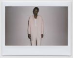 Une personne se tient debout devant un fond uni, vêtue d'un élégant costume rose clair avec une longue veste et un pantalon ajusté, associé à un haut blanc. Prise à Paris par Thomas Duclert, la photo est dotée d'une bordure Polaroid, ce qui lui confère un aspect vintage.