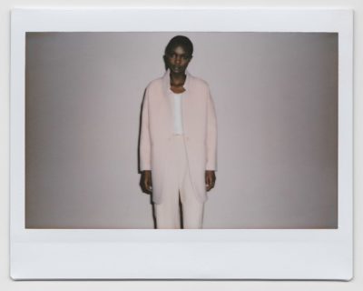 Une personne se tient debout devant un fond uni, vêtue d'un élégant costume rose clair avec une longue veste et un pantalon ajusté, associé à un haut blanc. Prise à Paris par Thomas Duclert, la photo est dotée d'une bordure Polaroid, ce qui lui confère un aspect vintage.