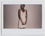 Une mannequin se tient debout sur un fond uni, le regard pensif. Elle porte une robe rose pâle sans manches et a les cheveux courts. Prise par un photographe talentueux, l'image présente une bordure de style Polaroïd rappelant la mode parisienne.