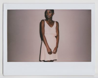 Une mannequin se tient debout sur un fond uni, le regard pensif. Elle porte une robe rose pâle sans manches et a les cheveux courts. Prise par un photographe talentueux, l'image présente une bordure de style Polaroïd rappelant la mode parisienne.