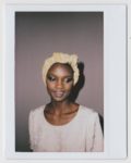 Une mannequin à la peau foncée, coiffée d'un foulard à motifs clairs et d'une tenue assortie, se tient debout sur un fond uni. Son expression est sereine, ses yeux légèrement baissés. La photo, qui rappelle la mode parisienne, présente une bordure de style Polaroïd qui capture le talent artistique du photographe.