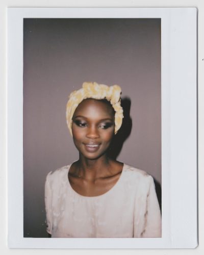 Une mannequin à la peau foncée, coiffée d'un foulard à motifs clairs et d'une tenue assortie, se tient debout sur un fond uni. Son expression est sereine, ses yeux légèrement baissés. La photo, qui rappelle la mode parisienne, présente une bordure de style Polaroïd qui capture le talent artistique du photographe.
