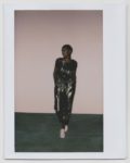 Une personne se tient devant un mur uni, vêtue d'une combinaison sombre et brillante et de chaussures blanches. Photographiée par le photographe Thomas Duclert, elle a les cheveux courts et regarde sur le côté, les mains dans les poches. Le décor dégage une esthétique simple et minimaliste qui rappelle le style parisien.