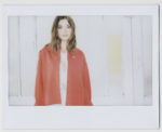 Une mannequin aux cheveux ondulés se tient devant un fond en bois blanc. Elle porte une chemise rose sous un cardigan ample rouge vif. L'image, capturée par un photographe talentueux à Paris, a un effet doux et rêveur avec un éclairage doux projetant une lueur chaleureuse sur la scène.