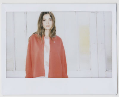 Une mannequin aux cheveux ondulés se tient devant un fond en bois blanc. Elle porte une chemise rose sous un cardigan ample rouge vif. L'image, capturée par un photographe talentueux à Paris, a un effet doux et rêveur avec un éclairage doux projetant une lueur chaleureuse sur la scène.