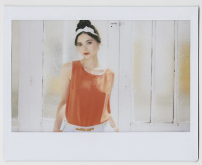 Une mannequin aux cheveux noirs attachés par un bandeau blanc porte un chemisier orange sans manches. Elle se tient devant une fenêtre au verre dépoli, regardant l'objectif avec une expression légèrement sérieuse, capturée par le photographe Thomas Duclert à Paris.
