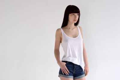 Une mannequin aux cheveux longs, raides et noirs, avec une frange, se tient debout sur un fond blanc. Elle porte un débardeur blanc ample et un short en jean, une main posée sur sa hanche et le regard légèrement sur le côté. Captured par le talentueux photographe Thomas Duclert, son élégance décontractée transparaît.