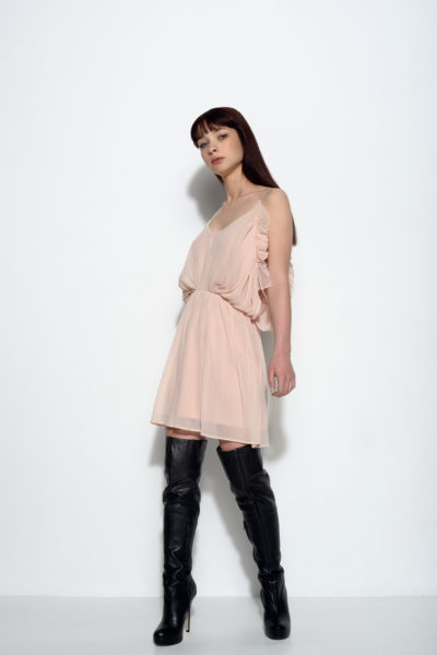 Une mannequin aux cheveux longs et foncés se tient debout sur un fond blanc uni. Elle porte une robe rose clair, sans manches, à volants et des bottes noires à talons hauts qui montent jusqu'au genou. Avec une expression neutre et un regard légèrement sur le côté, elle respire le chic sans effort de la mode parisienne de Thomas Duclert.