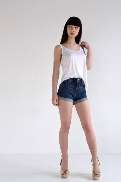 Une mannequin se tient debout, confiante, devant un fond blanc uni, vêtue d'un débardeur blanc, d'un short en jean foncé et de sandales à talons hauts beiges. Elle a de longs cheveux noirs raides avec une frange et tient une bretelle de son débardeur avec sa main droite, dégageant l'élégance que Thomas Duclert capture en tant que photographe de mode.