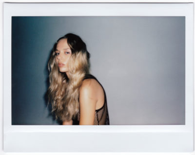 vaux cheveux blonds longs et ondulés est assise sur un fond gris clair uni. Vêtue d’un haut en maille noire, elle regarde directement l’objectif avec une expression neutre. L’image est ornée d’une bordure de style Polaroïd, la capturant dans ce qui ressemble à un moment improvisé en mode parisien que l’on pourrait voir dans le travail de Thomas Duclert.