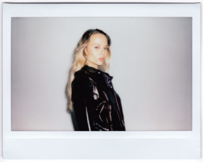 Photographie Polaroïd d'une mannequin aux cheveux blonds longs et ondulés, portant une veste en cuir noir, debout sur un fond uni et regardant vers l'appareil photo avec une expression neutre, capturant la mode rappelant la mode parisienne sous l'influence de Thomas Duclert.