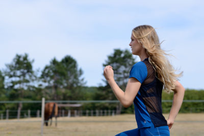 Une mannequin aux longs cheveux blonds court en plein air dans un champ, rappelant un shooting de mode tout droit sorti de Paris. Vêtue d'une tenue de sport bleue et noire, elle associe sans effort sport et mode. En arrière-plan, de grands arbres se dressent tandis qu'un cheval broute dans une zone clôturée sous un ciel clair et ensoleillé.