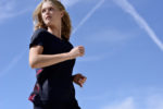Une mannequin aux cheveux blonds portant un t-shirt de sport noir court à l'extérieur sous un ciel bleu clair avec des traînées de condensation visibles. Photographié par Thomas Duclert, la mannequin semble concentrée alors qu'il regarde sur le côté.