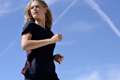Une mannequin aux cheveux blonds portant un t-shirt de sport noir court à l'extérieur sous un ciel bleu clair avec des traînées de condensation visibles. Photographié par Thomas Duclert, la mannequin semble concentrée alors qu'il regarde sur le côté.