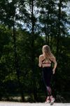 Une mannequin aux longs cheveux blonds court sur un sentier en forêt. Elle porte un soutien-gorge de sport noir, un legging noir à motifs rouges et des chaussures de course roses. Le photographe Thomas Duclert capture sa foulée déterminée au milieu des arbres au feuillage dense sous un ciel clair de Paris.