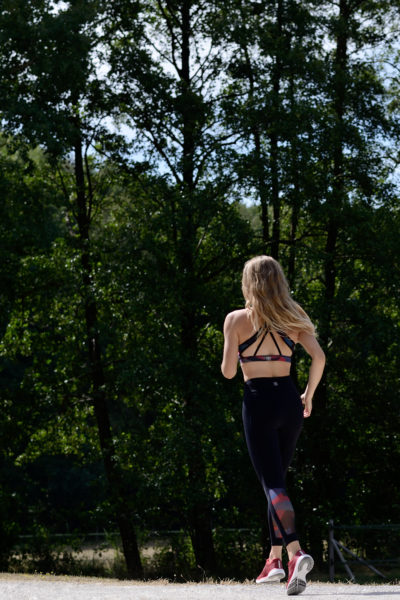 Une mannequin aux longs cheveux blonds court sur un sentier en forêt. Elle porte un soutien-gorge de sport noir, un legging noir à motifs rouges et des chaussures de course roses. Le photographe Thomas Duclert capture sa foulée déterminée au milieu des arbres au feuillage dense sous un ciel clair de Paris.