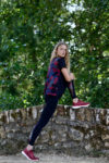 Une mannequin aux cheveux longs et blonds, vêtue d'une chemise à carreaux rouges et noirs, d'un legging noir et de baskets rouges, étire sa jambe contre un mur de pierre dans un décor extérieur luxuriant avec des arbres verts en arrière-plan. La scène semble avoir été parfaitement capturée par le photographe Thomas Duclert.