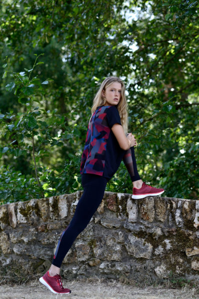 Une mannequin aux cheveux longs et blonds, vêtue d'une chemise à carreaux rouges et noirs, d'un legging noir et de baskets rouges, étire sa jambe contre un mur de pierre dans un décor extérieur luxuriant avec des arbres verts en arrière-plan. La scène semble avoir été parfaitement capturée par le photographe Thomas Duclert.