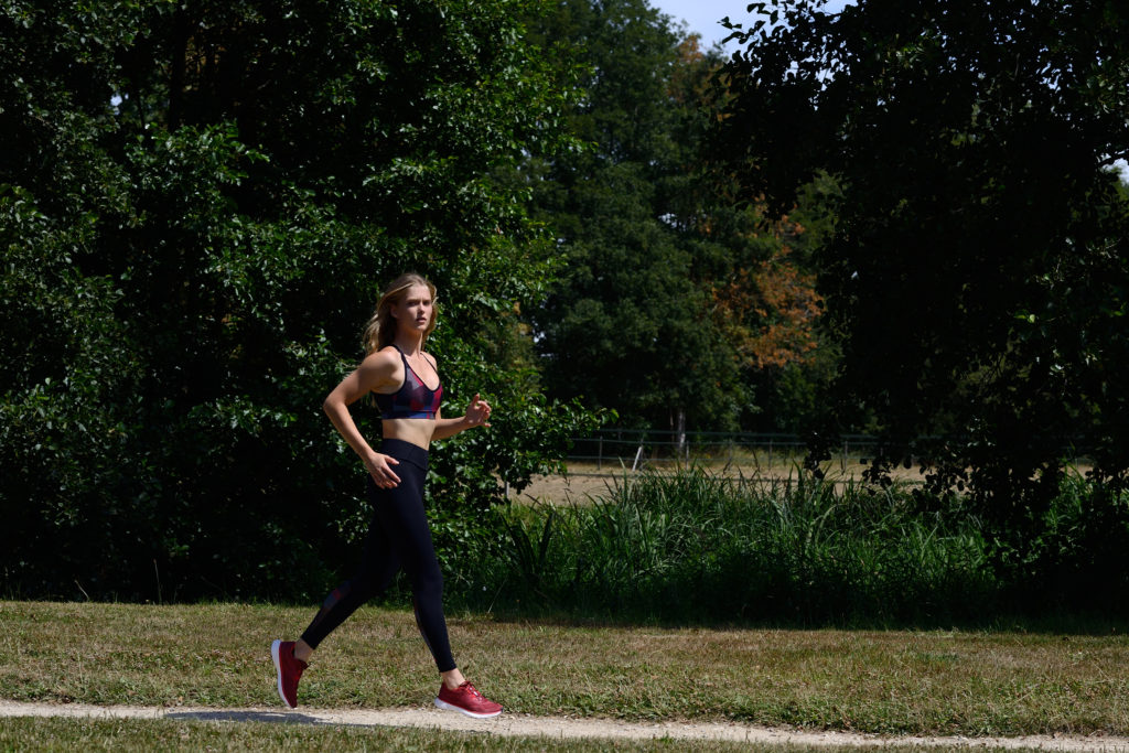 Une mannequin portant un soutien-gorge de sport, un legging et des baskets rouges fait du jogging sur un chemin de gravier dans un parc. Photographiée par Thomas Duclert, elle est entourée d'arbres verts luxuriants et d'herbe sous un ciel bleu clair. Elle semble concentrée et déterminée.