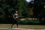 Une mannequin portant un soutien-gorge de sport, un legging et des baskets rouges fait du jogging sur un chemin de gravier dans un parc. Photographiée par Thomas Duclert, elle est entourée d'arbres verts luxuriants et d'herbe sous un ciel bleu clair. Elle semble concentrée et déterminée.
