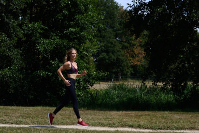 Une mannequin portant un soutien-gorge de sport, un legging et des baskets rouges fait du jogging sur un chemin de gravier dans un parc. Photographiée par Thomas Duclert, elle est entourée d'arbres verts luxuriants et d'herbe sous un ciel bleu clair. Elle semble concentrée et déterminée.
