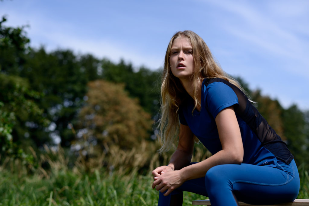 Une mannequin aux longs cheveux blonds, vêtue d'une tenue de sport bleue, est assise à l'extérieur sur un banc, pensive. Elle regarde légèrement vers le haut, son expression contemplative. L'arrière-plan présente des arbres verts et de l'herbe sous un ciel bleu, rappelant les journées tranquilles à Paris.