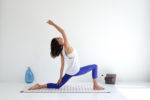 Une mannequin en haut blanc et legging bleu exécute une posture de yoga sur un tapis à l'intérieur, rappelant une séance photo sereine de mode parisienne. Elle est en position de fente basse avec sa jambe gauche en avant, sa jambe droite tendue vers l'arrière et son bras droit tendu au-dessus de sa tête. Un vase bleu et des blocs de yoga empilés sont en arrière-plan.