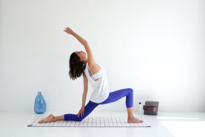 Une mannequin en haut blanc et legging bleu exécute une posture de yoga sur un tapis à l'intérieur, rappelant une séance photo sereine de mode parisienne. Elle est en position de fente basse avec sa jambe gauche en avant, sa jambe droite tendue vers l'arrière et son bras droit tendu au-dessus de sa tête. Un vase bleu et des blocs de yoga empilés sont en arrière-plan.