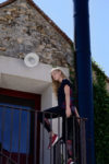 Une mannequin aux longs cheveux blonds escalade une balustrade en métal à l'extérieur d'un immeuble en pierre à Paris. elle est vêtue de vêtements sombres décontractés et de baskets rouges. Le bâtiment a une façade en pierre, un luminaire moderne au-dessus d'une porte rouge et du lierre vert grimpe le long du mur, dégageant un style décontracté rappelant l'esthétique de Thomas Duclert.