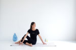 Une mannequin exécute une torsion de yoga assis sur un tapis à carreaux blancs dans une pièce minimaliste avec un fond blanc, évoquant le style d'un photographe de mode à Paris. Elle porte une tenue noire et est positionnée sur le côté droit, avec un vase bleu et deux bougies colorées en arrière-plan.