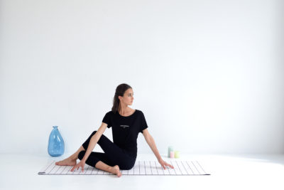 Une mannequin exécute une torsion de yoga assis sur un tapis à carreaux blancs dans une pièce minimaliste avec un fond blanc, évoquant le style d'un photographe de mode à Paris. Elle porte une tenue noire et est positionnée sur le côté droit, avec un vase bleu et deux bougies colorées en arrière-plan.