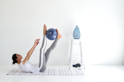Une mannequin en tenue de sport est allongée sur le dos sur un tapis de yoga, soulevant un ballon d'exercice bleu avec ses pieds et tendant les bras dans une pièce lumineuse et minimaliste. Capturée par Thomas Duclert, la scène comprend un tabouret blanc avec un vase bleu et des haltères en arrière-plan.
