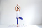 Une mannequin se tient debout sur une jambe dans une posture de yoga en forme d'arbre sur un tapis rayé. Elle porte un débardeur blanc et un legging bleu, les mains jointes au-dessus de la tête. L'arrière-plan est un mur blanc uni, avec quelques accessoires de yoga sur le sol. Ce moment serein a été capturé avec brio par le photographe parisien Thomas Duclert.