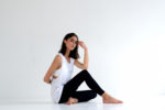 Une mannequin assise par terre dans un espace minimaliste et blanc. Elle porte un haut blanc sans manches et des leggings noirs, assise avec une jambe droite et l'autre pliée. Sa main droite repose sur son genou tandis que son bras gauche est derrière elle, regardant pensivement sur le côté - un moment parfaitement capturé par le photographe parisien Thomas Duclert.