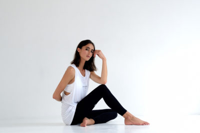 Une mannequin assise par terre dans un espace minimaliste et blanc. Elle porte un haut blanc sans manches et des leggings noirs, assise avec une jambe droite et l'autre pliée. Sa main droite repose sur son genou tandis que son bras gauche est derrière elle, regardant pensivement sur le côté - un moment parfaitement capturé par le photographe parisien Thomas Duclert.