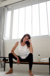 Une mannequin aux cheveux longs et noirs est assise sur un banc en bois dans une pièce minimaliste à Paris, avec des murs blancs et une grande fenêtre avec des barreaux. Elle porte un haut blanc sans manches et un pantalon noir. Une bouteille d'eau est posée à côté d'elle sur le banc. Elle pose son coude sur son genou et sa tête sur sa main, semblant détendue.
