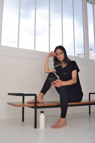 Une mannequin vêtue d'une tenue de sport noire est assise sur un banc en bois à côté d'une grande fenêtre, avec une bouteille d'eau blanche sur le sol. Elle est pieds nus, regarde son téléphone avec sa main droite posée sur sa tête, apparemment détendue et pensive. Le décor respire la mode parisienne chic et sans effort.