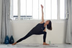 Une mannequin en tenue de sport noire exécute une posture de yoga en angle latéral sur un tapis à l'intérieur. La personne se penche en avant sur une jambe tout en étendant le bras opposé vers le haut, l'autre bras reposant sur la jambe avant. De grandes fenêtres à l'arrière-plan laissent entrer la lumière naturelle, capturant parfaitement un moment serein digne de l'objectif de Thomas Duclert.