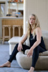 Une mannequin aux longs cheveux blonds est assise sur un pouf beige dans un salon moderne. Elle porte un haut sans manches noir et des leggings noirs, regardant pensivement la caméra. Capturée par Thomas Duclert, la pièce présente des murs carrelés de couleur claire et des meubles en bois en arrière-plan.