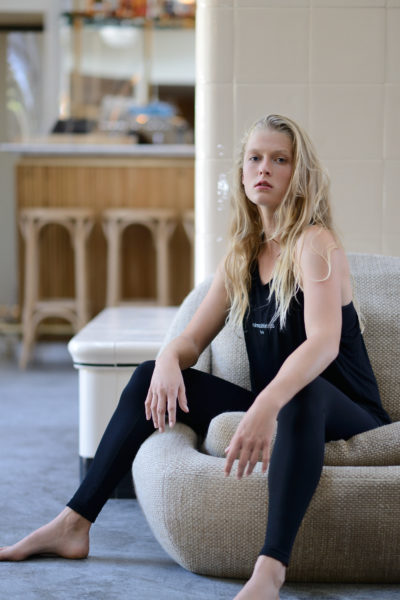 Une mannequin aux longs cheveux blonds est assise sur un pouf beige dans un salon moderne. Elle porte un haut sans manches noir et des leggings noirs, regardant pensivement la caméra. Capturée par Thomas Duclert, la pièce présente des murs carrelés de couleur claire et des meubles en bois en arrière-plan.
