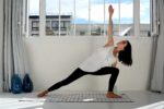 Une mannequin en débardeur blanc et leggings noirs pratique le yoga en intérieur, en effectuant une pose allongée sur un tapis à carreaux. La pièce, photographiée par le photographe parisien Thomas Duclert, a des murs blancs, une grande fenêtre avec vue sur le paysage urbain et deux vases bleus sur le sol.
