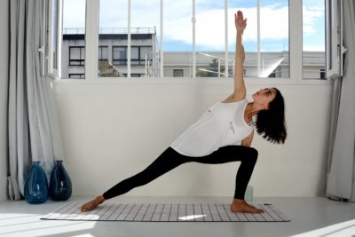 Une mannequin en débardeur blanc et leggings noirs pratique le yoga en intérieur, en effectuant une pose allongée sur un tapis à carreaux. La pièce, photographiée par le photographe parisien Thomas Duclert, a des murs blancs, une grande fenêtre avec vue sur le paysage urbain et deux vases bleus sur le sol.