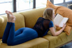 Une mannequin aux cheveux longs est allongée sur le ventre sur un canapé jaune moutarde, en train de lire un livre. Elle porte un legging bleu et un haut en maille à manches courtes. Avec deux gros coussins derrière elle et la lumière du soleil qui entre par la fenêtre, on se croirait dans une scène tout droit sortie de Paris, à la manière de Thomas Duclert.