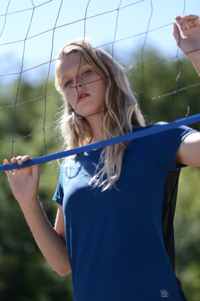 Une mannequin aux longs cheveux blonds, vêtue d'une chemise bleue, pose devant un filet de volley-ball extérieur. Elle regarde directement l'objectif, une main tenant le filet et l'autre agrippant le poteau, capturée dans une photographie de Thomas Duclert sur un fond flou de verdure.